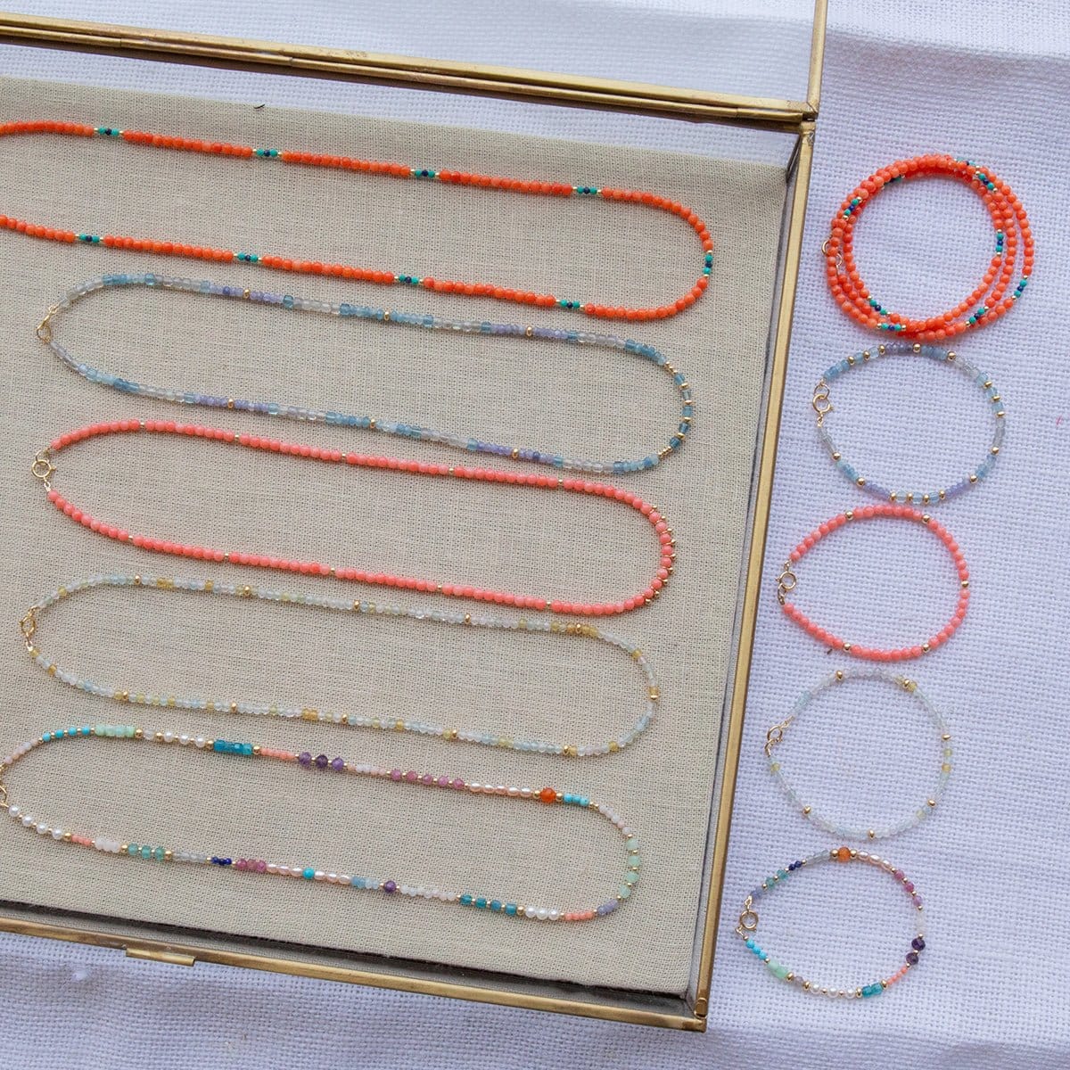 Endless Summer Necklaces and Bracelets (Calypso, Blue Lagoon, Mykonos, Saint-Tropez and Capri), with the necklaces laid in a clear tray on beige fabric and the bracelets laid next to the tray on a white fabric background.