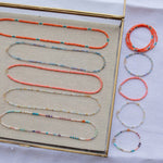 Endless Summer Necklaces and Bracelets (Calypso, Blue Lagoon, Mykonos, Saint-Tropez and Capri), with the necklaces laid in a clear tray on beige fabric and the bracelets laid next to the tray on a white fabric background.