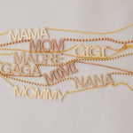 A MAMA Necklace, GIGI Necklace, Pavé MADRE Necklace, Pavé GAGA Necklace, Pavé NANA Necklace, and a MOMMY Necklace in yellow gold, along with Pavé MOM and MIMI Necklaces in rose gold, laid on an off-white background.