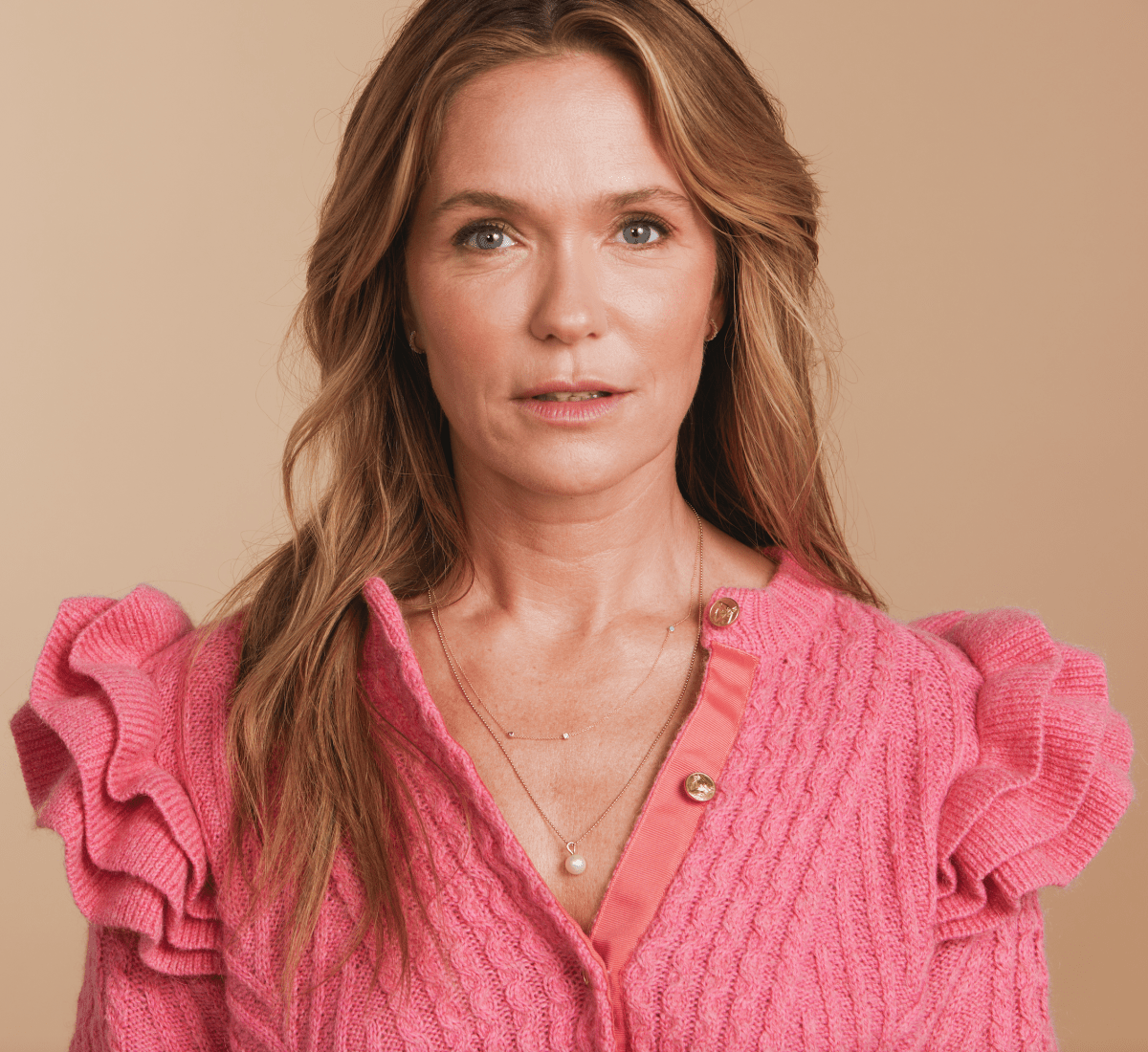 A woman wearing a Custom Gold Necklace with three birthstone charms and a Perla Necklace, styled in a pink sweater with ruffled sleeves against a beige background. 