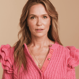A woman wearing a Custom Gold Necklace with three birthstone charms and a Perla Necklace, styled in a pink sweater with ruffled sleeves against a beige background. 