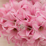 small nameplate necklace in lowercase 'otis' in white gold on a pink flower bouquet 