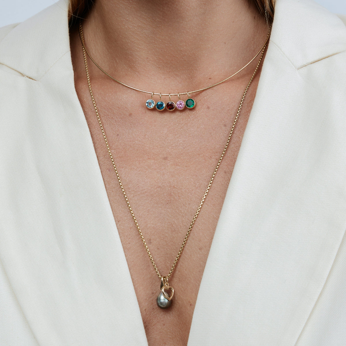 Close-up of a woman wearing an Omega Chain with Color Pop Pendants, and a Box Chain with a Cut Out Heart Charm with a Baroque Pearl Pendant, styled in a white blazer. 