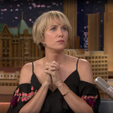 Kristen Wiig wearing an Omega Chain with several other gold jewellery pieces sitting on a talk show set with a cityscape background. 