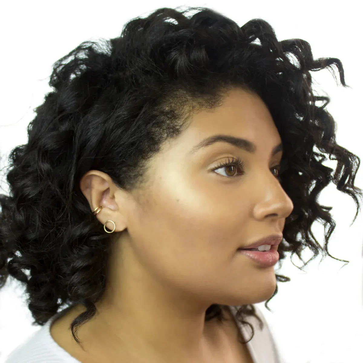 A woman wearing a Double Ear Cuff and Open Eclipse Earring - Small in yellow gold, with black curly hair looking to the side, against a white background. 