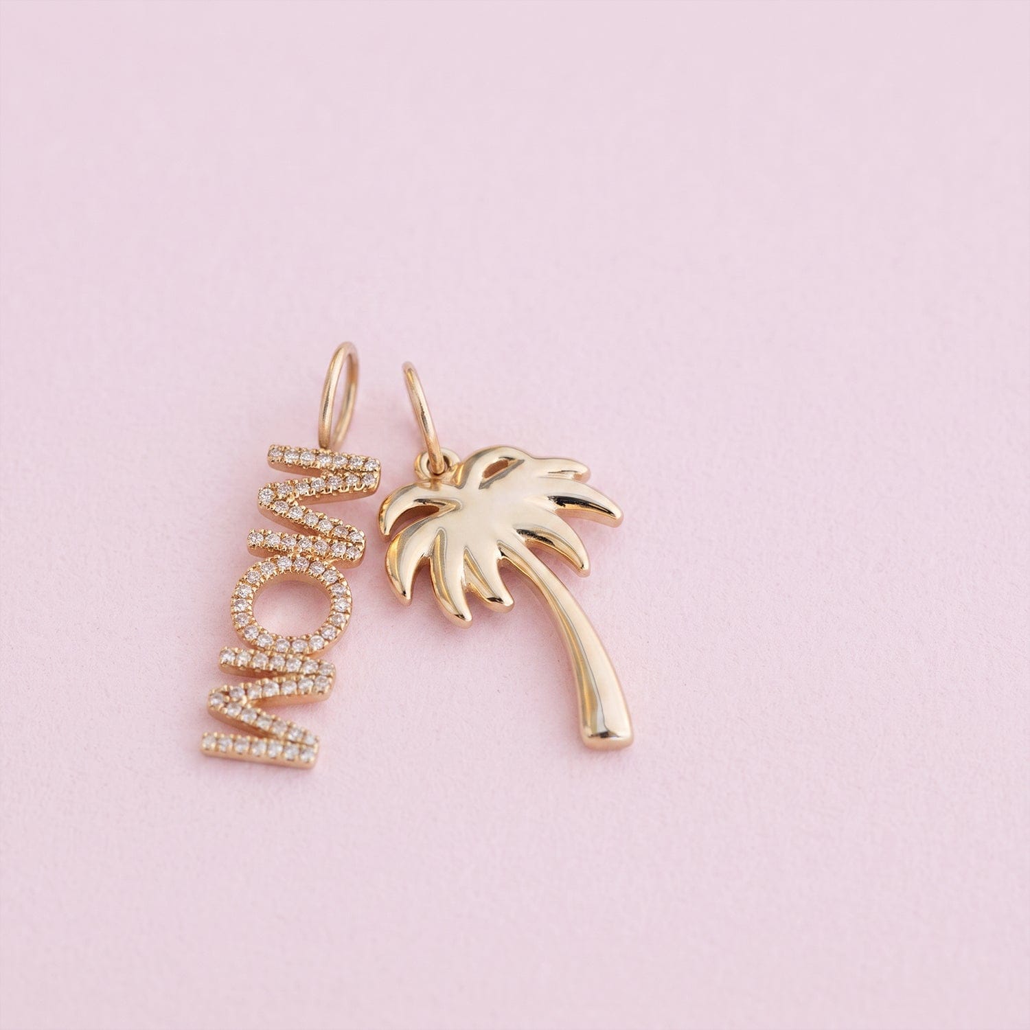 A pair of gold-toned jewelry charms on a soft pink background. One charm is a vertical script word 'MOM' set with small clear stones, and the other is a palm tree silhouette in polished gold.