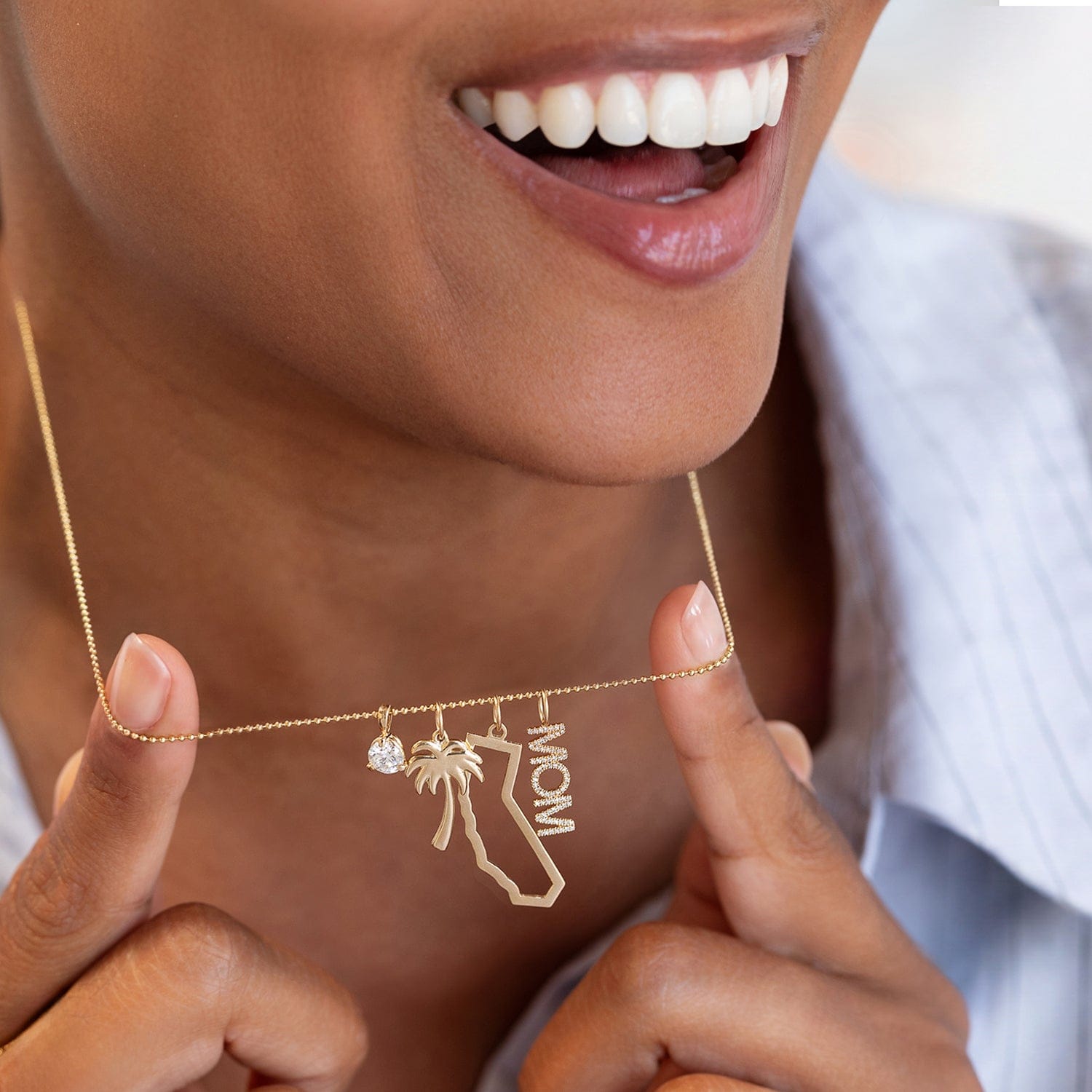 A close-up lifestyle shot of a woman holding a delicate gold necklace with charms. The necklace features a small clear gemstone, a palm tree, a California state silhouette, and the word 'MOM'.