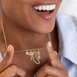 A close-up lifestyle shot of a woman holding a delicate gold necklace with charms. The necklace features a small clear gemstone, a palm tree, a California state silhouette, and the word 'MOM'.