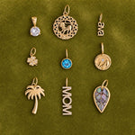 Nine gold-toned jewelry pendants arranged in a 3x3 grid on a textured olive green background. The pendants include a round clear gemstone, a circular filigree pendant with a lion motif, a script nameplate reading 'ava', a small flower with clear stones, a round blue gemstone pendant, a circular medallion with a sun motif, a palm tree pendant, a vertical 'MOM' nameplate with clear stones, and a leaf-shaped pendant with a purple and blue inlay.