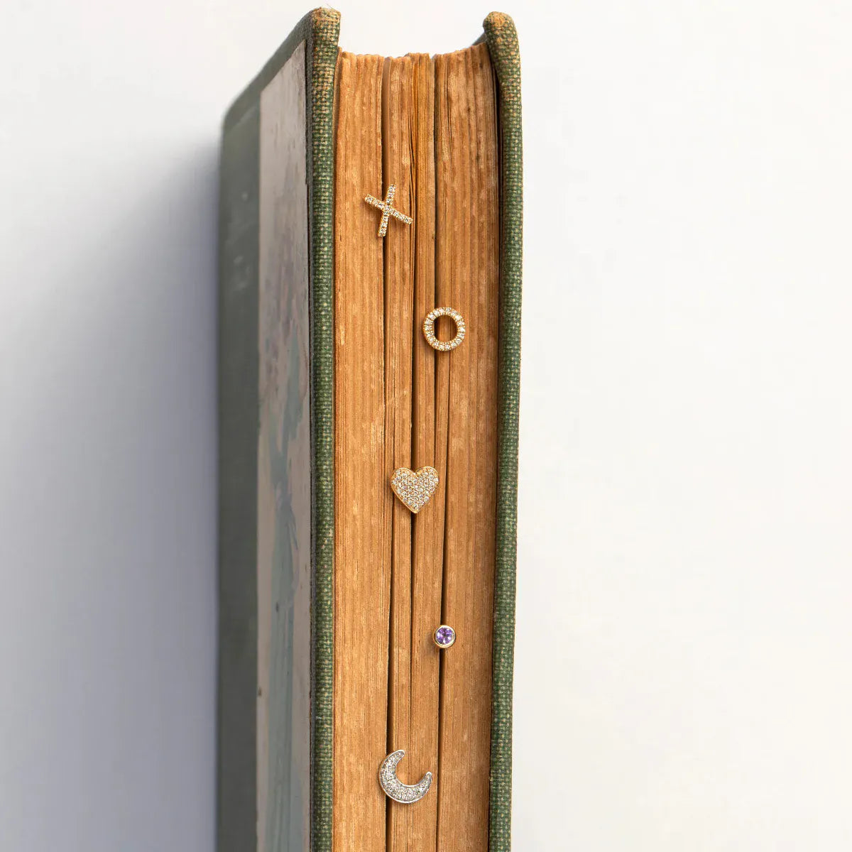 Pavé Charm Studs - Heart and Moon, a Birthstone Stud and two 14K Gold Pave Letter Studs in X and O displayed on top of a book against a white background. 