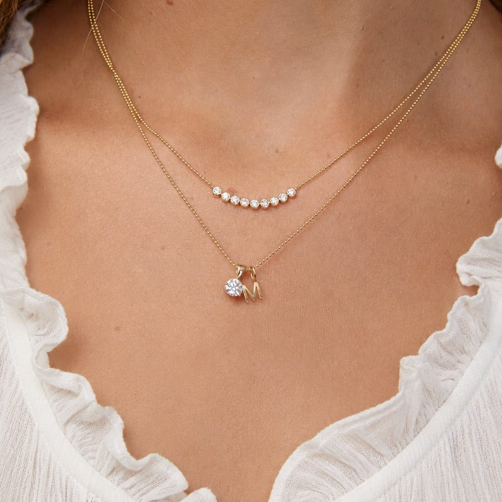 Gold necklace with a letter 'M' and pave cross pendant on a person wearing a white top.