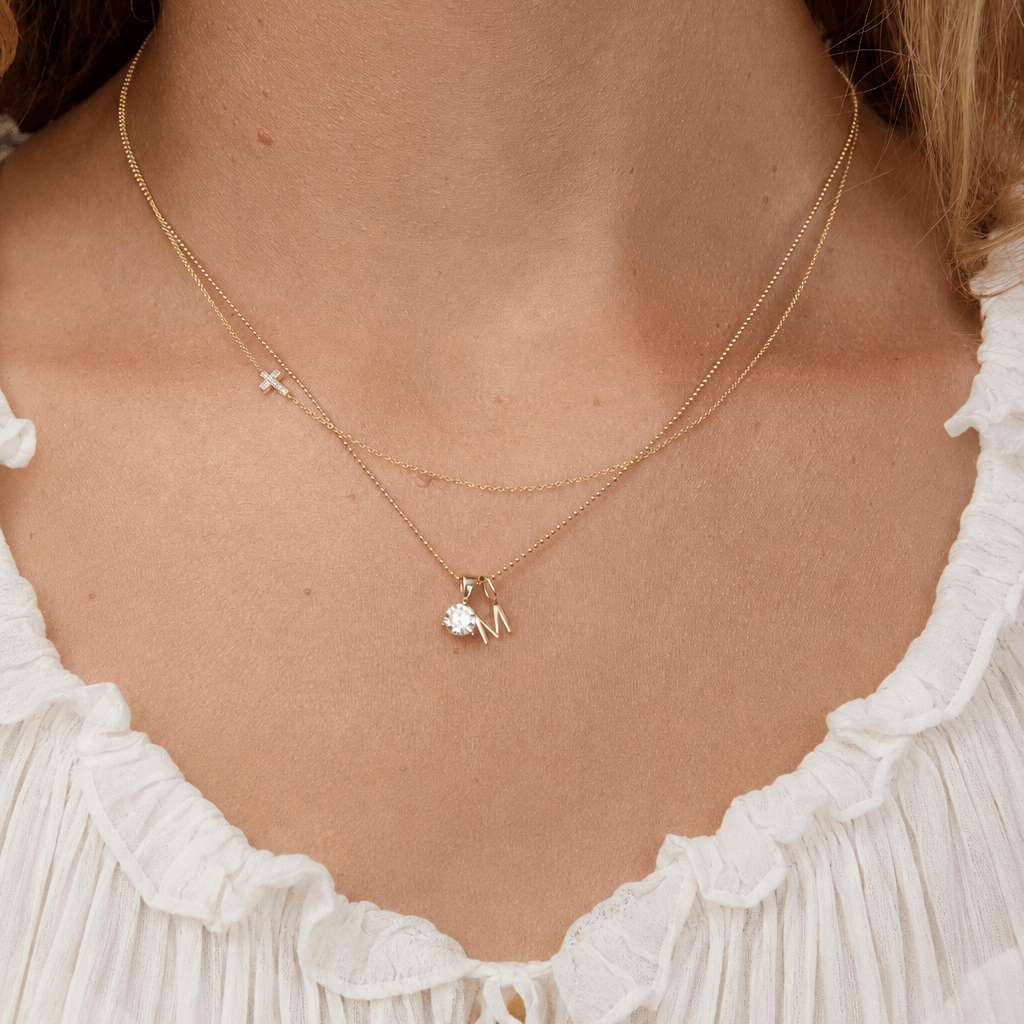 Gold necklace with a letter 'M' pendant on a person wearing a white top.