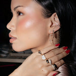 Close-up of a woman wearing a Diamond Fizz Earring, Highball Diamond Chain Earring, two Pave Ear Cuffs, a Pinky Promise Ring, Love on the Rocks Ring, Make it a Double Ring, Cosmo Ring and Martini Ring with red nails, with a dark background. 