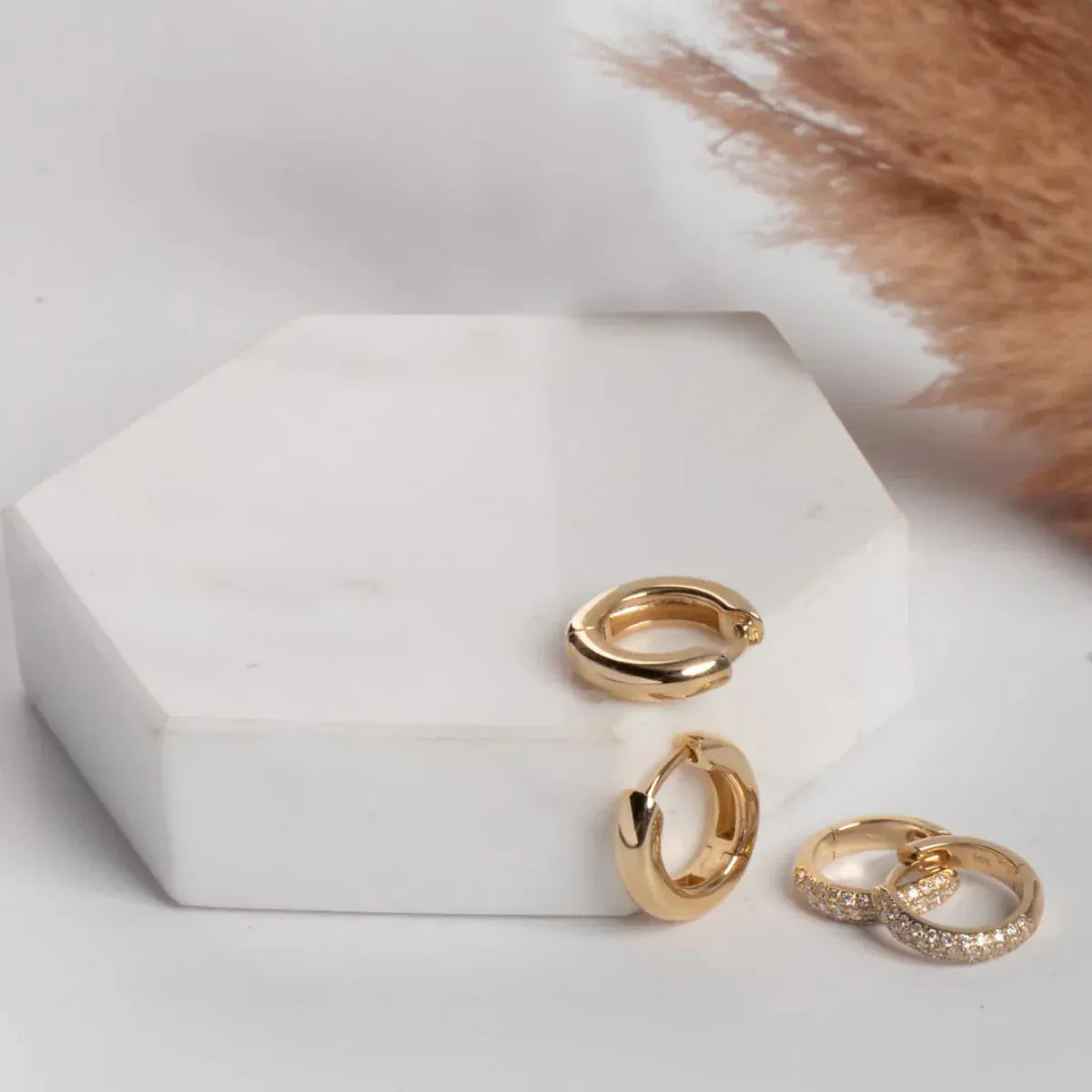 A pair of Gold Huggies and Pave Huggies on a white hexagonal stone and surface, with a soft brown piece of fabric in the top corner.