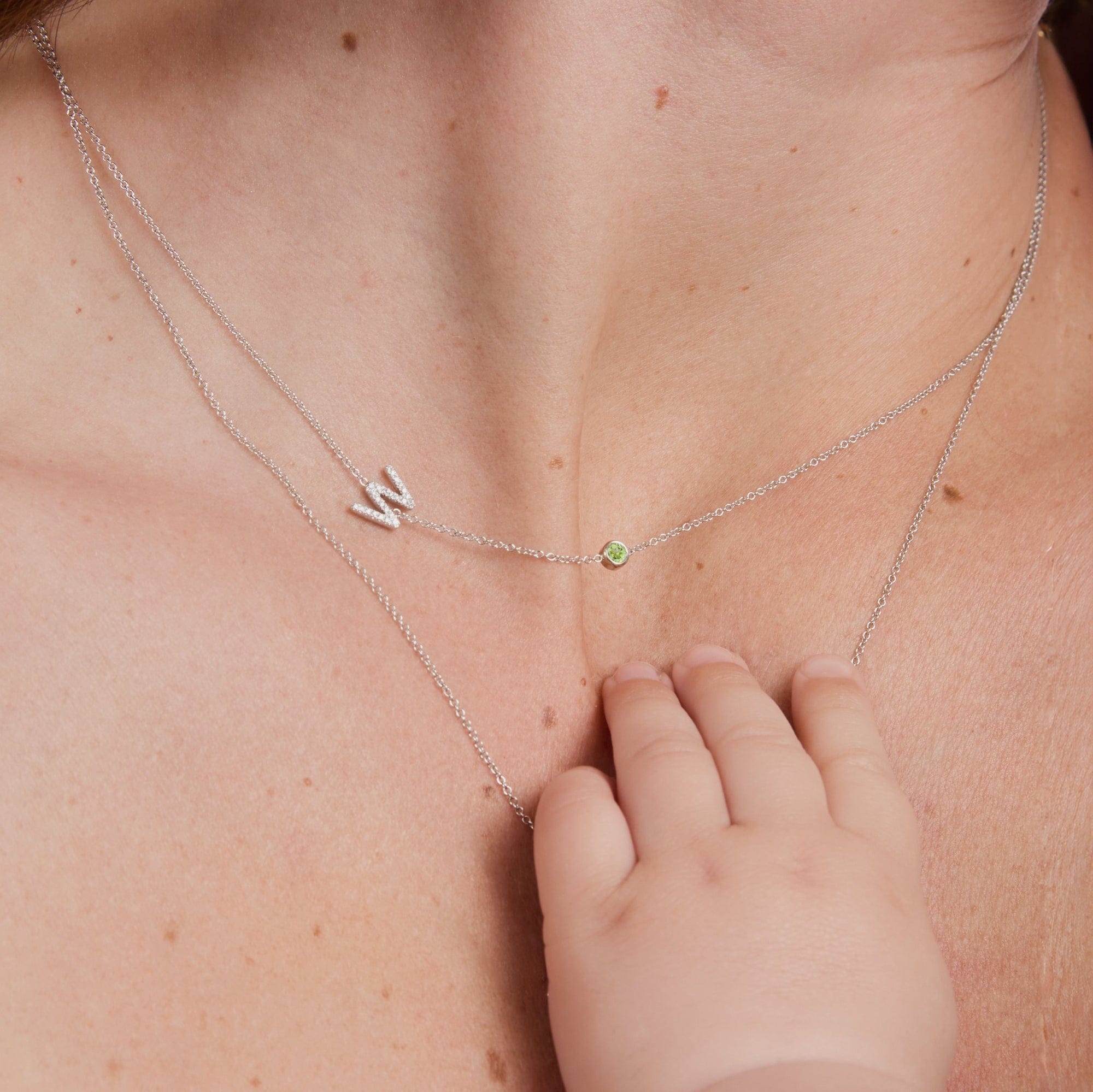 A close-up of a delicate double-layered white gold necklace worn on a baby's chest, featuring a letter 'W' pendant and a small green gemstone, with a baby's hand gently resting on the chain.