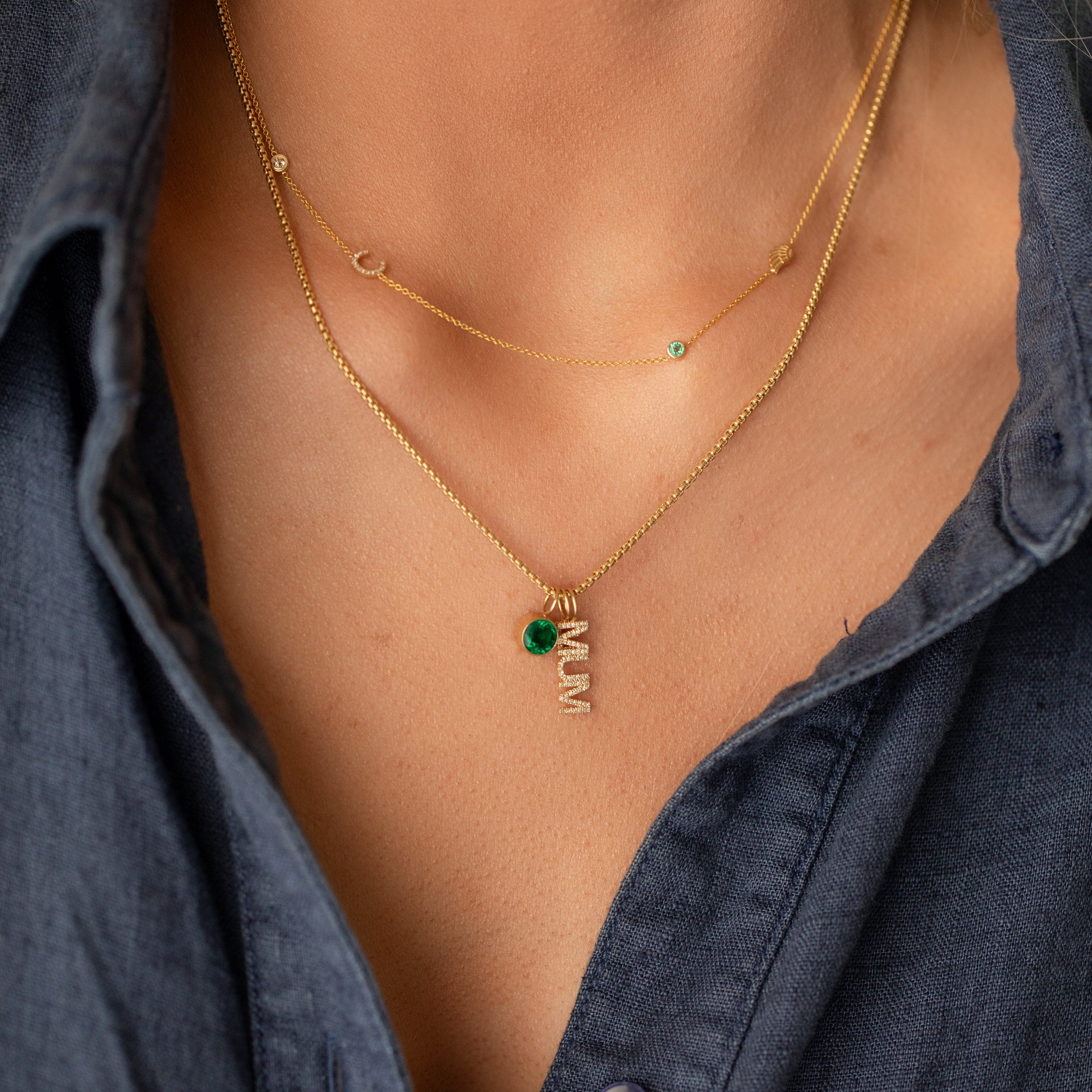 Gold necklace with a green gemstone pendant and 'MUM' pendant on a person wearing a dark blue shirt.