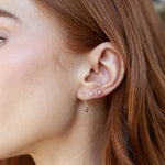 A close-up of a woman's ear wearing two Birthstone Studs (Diamond and Pearl) and a Gold Safety Pin Earring, with orange hair. 