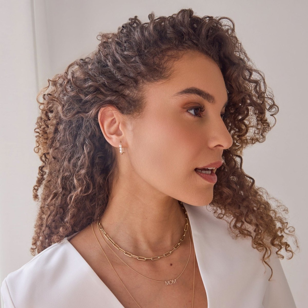 A woman wearing a Tres Pearl Earring, Bold Link Chain, MOM Necklace and Perla Necklace, with brown curly hair and a white top against a white background. 