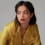 A brunette woman wearing a Perla Necklace, a 20th Anniversary Leaf Necklace and Sleek & Chic Hoops - Small, styled in a mustard yellow jacket against a light grey background. 