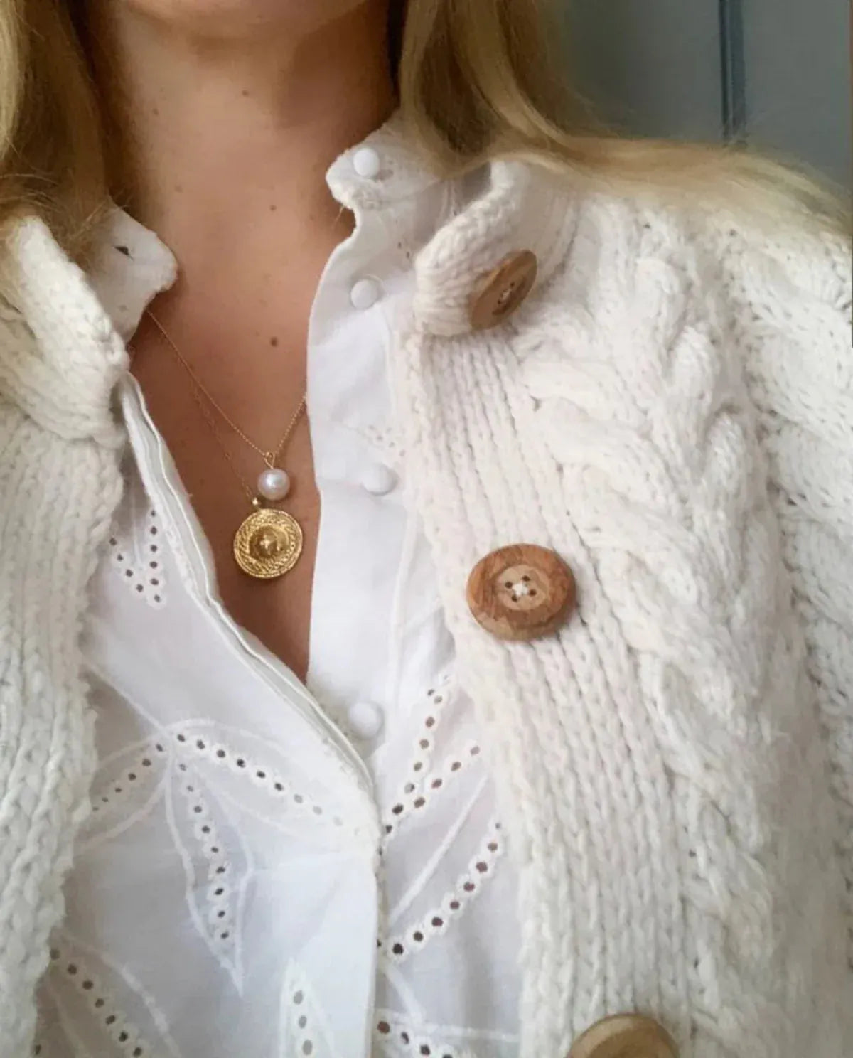 Close-up of a blonde woman's décolletage, wearing a Perla Necklace paired with another gold necklace, styled in a white shirt and cable knit cardigan with wooden buttons. 