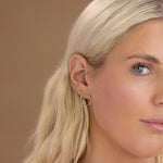 Close-up a Birthstone Stud - Peridot (August) and a Open Eclipse Earring - Large in a blonde woman's ear against a light brown background. 