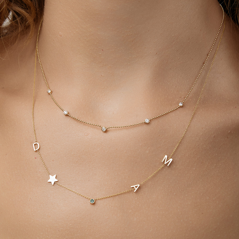 Gold necklace with letter charms on a person's neck layered with a necklace that has five diamonds on it.