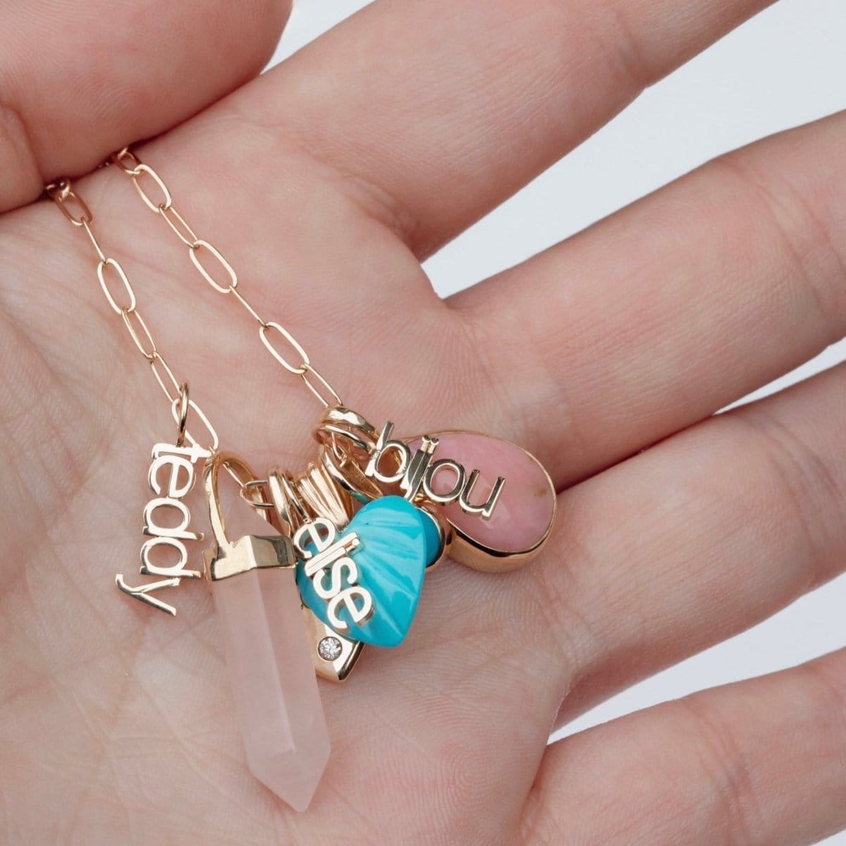 A hand holding an Element Short Link Chain with several charms, including Petite Nameplate Pendants 'bijou', 'teddy' and 'elise', an Etched Turquoise Heart Charm, a Balance Crystal Charm, an Engraved Heart Charm and an Engraved Heart Charm. 