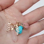 A hand holding an Element Short Link Chain with several charms, including Petite Nameplate Pendants 'bijou', 'teddy' and 'elise', an Etched Turquoise Heart Charm, a Balance Crystal Charm, an Engraved Heart Charm and an Engraved Heart Charm. 