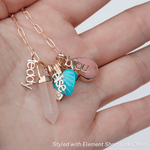 A hand holding an Element Short Link Chain with several charms, including Petite Nameplate Pendants 'bijou', 'teddy' and 'elise', an Etched Turquoise Heart Charm, a Balance Crystal Charm, an Engraved Heart Charm and an Engraved Heart Charm. 