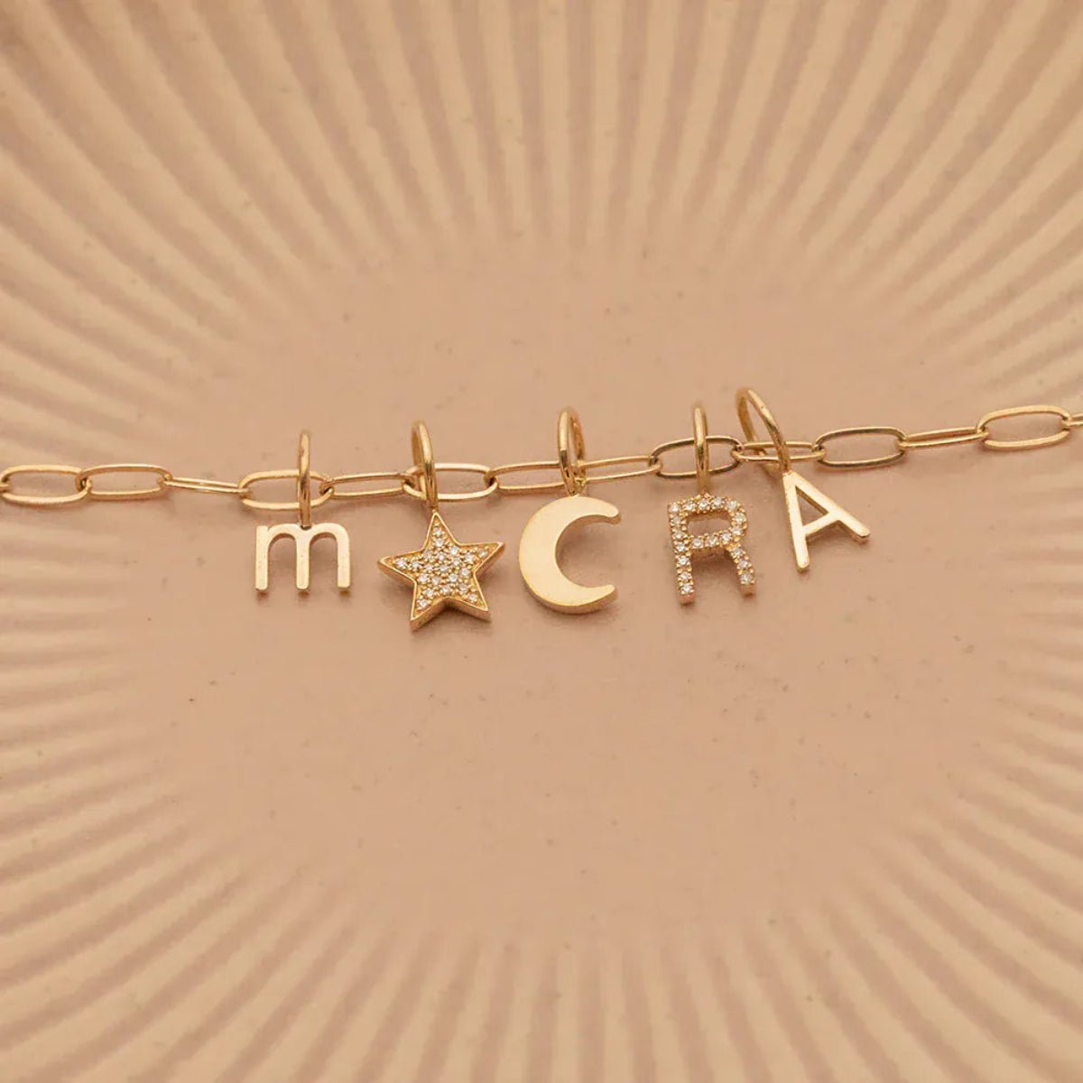 A Petite Letter Charm in m, a Gold Letter Charm in A, a Pavé Letter Charm in R, and Charm Pendants in Moon and Pavé Star on a Link Chain, all in yellow gold, on a beige background. 