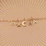 A Petite Letter Charm in m, a Gold Letter Charm in A, a Pavé Letter Charm in R, and Charm Pendants in Moon and Pavé Star on a Link Chain, all in yellow gold, on a beige background. 