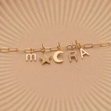 A Petite Letter Charm in m, a Gold Letter Charm in A, a Pavé Letter Charm in R, and Charm Pendants in Moon and Pavé Star on a Link Chain, all in yellow gold, on a beige background. 