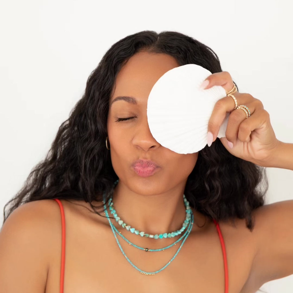 A video of a woman with curly black hair wearing Endless Summer Necklaces in Turqs + Caicos, Santorini, and Sea + Sky, along with several Bouquet Rings and a red strap top, holding a white shell over her face against a white background doing different poses. 