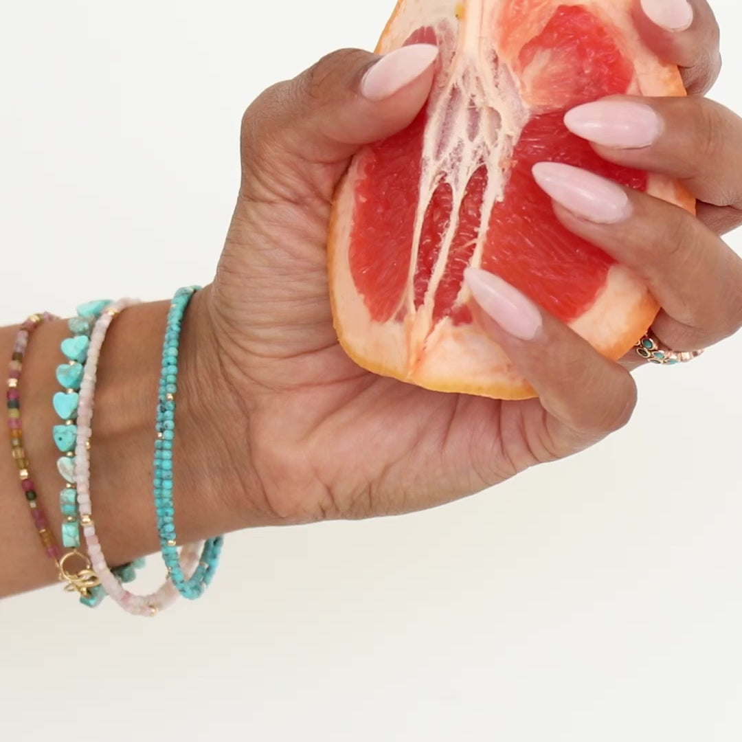A hand wearing Endless Summer Bracelets in Coral Reef, Sea + Sky, Pink Sands and Turqs + Caicos squeezing a grapefruit against a white background.