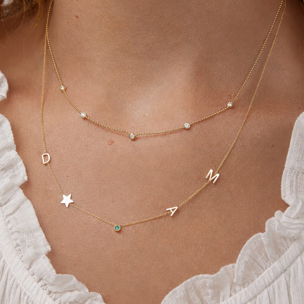 Gold necklace with letter charms on a person wearing a white lace top.