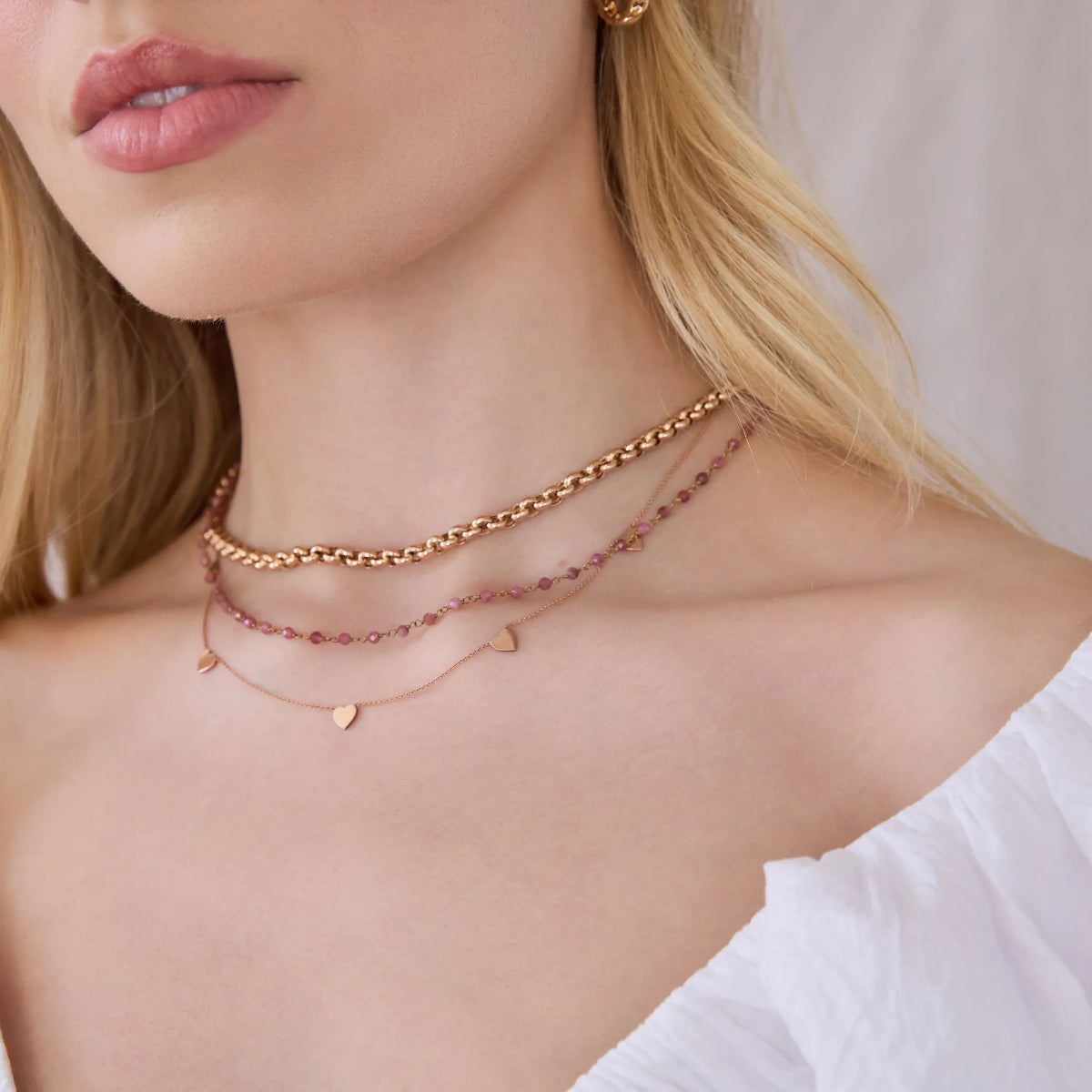Close-up of a blonde woman's neck wearing a rose gold Rolo Necklace, the Peony Bouquet Necklace and a 5 of Hearts Necklace, with a white top against a neutral background.