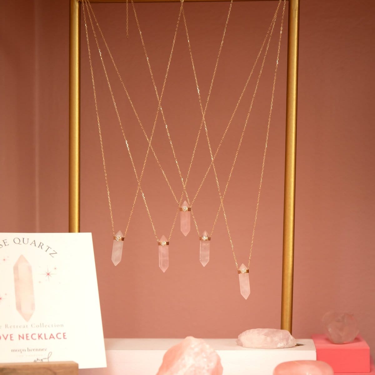 Several The Love Retreat Necklaces on a gold stand with rose quartz stones, against a pinky brown background. 