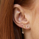 Close-up of a Pave Safety Pin Earring, 14K Gold Number Stud in 3 and a 14K Gold Pave Letter Stud in O in a woman's ear with her orange hair visible. 