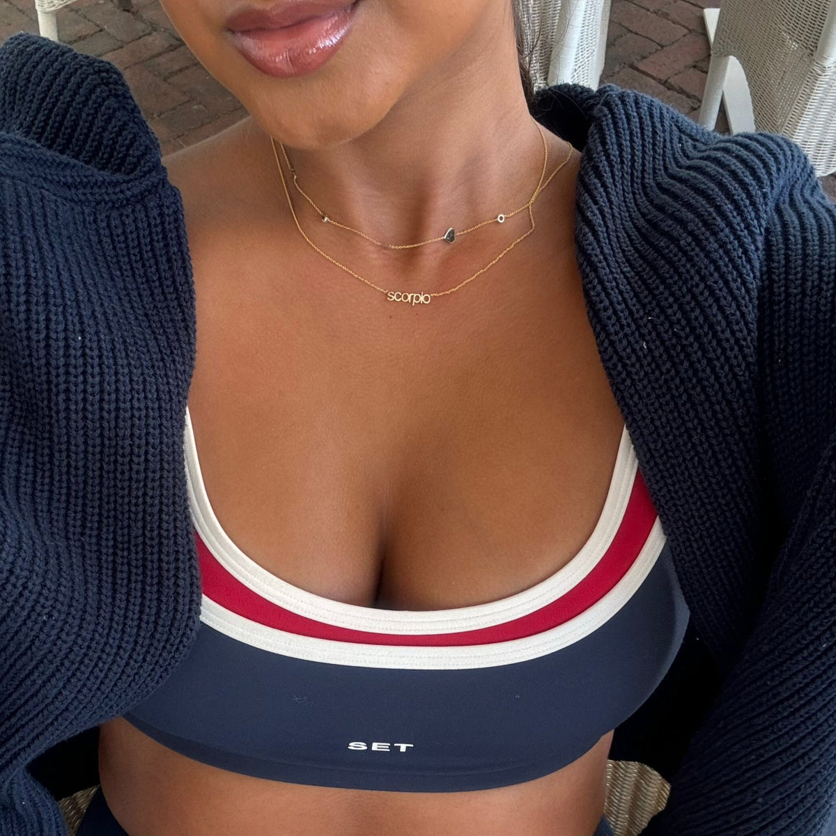 Person wearing a navy sports bra with red and white stripes and a navy cardigan and wearing a zodiac gold neclace and 4 charm gold necklace