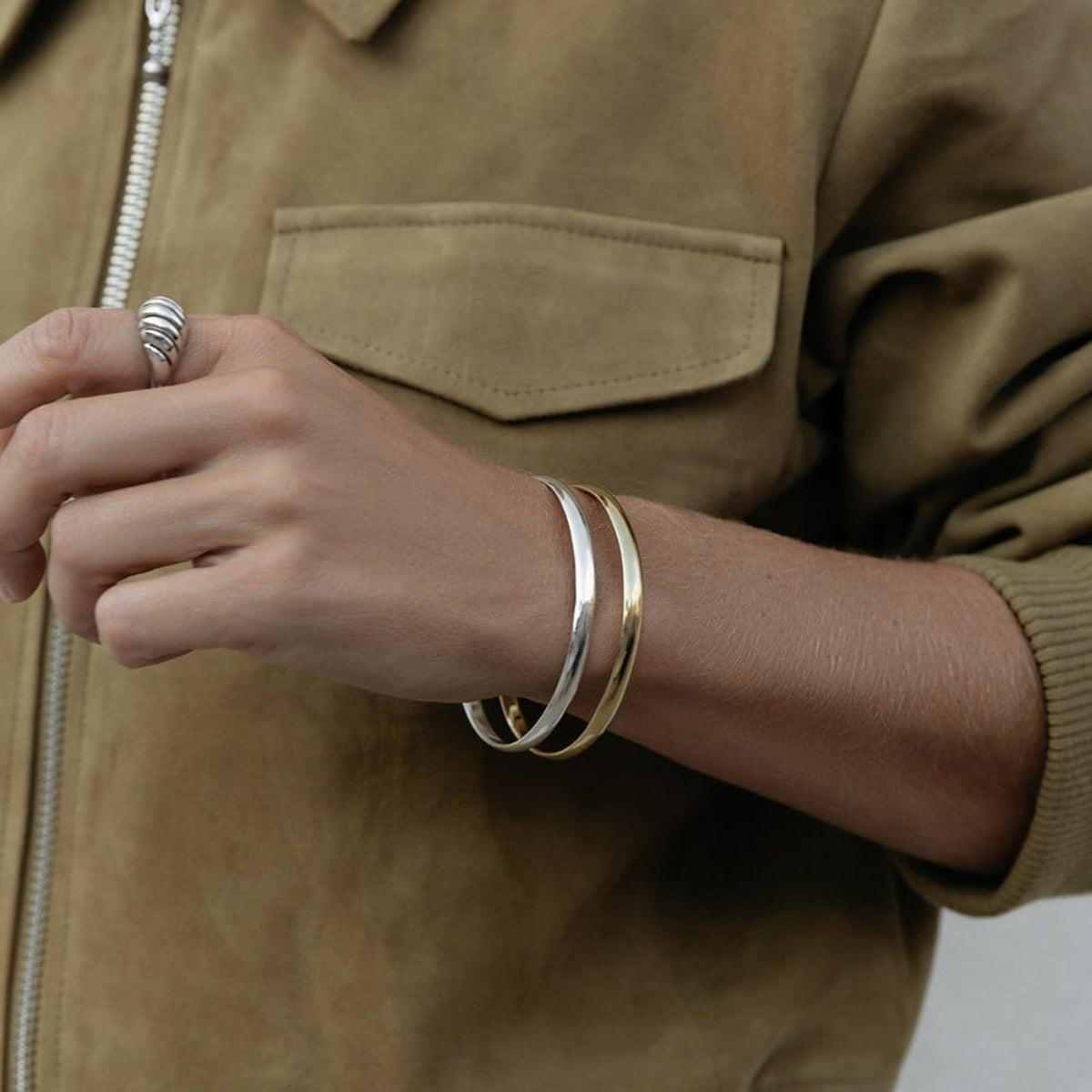A close-up of an arm wearing two For Keeps Bangle, one in gold and one in silver and a white gold Etched Paris Ring, styled in a khaki jacket. 