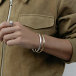 A close-up of an arm wearing two For Keeps Bangle, one in gold and one in silver and a white gold Etched Paris Ring, styled in a khaki jacket. 