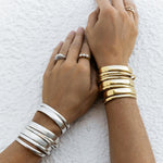 A close-up of arms wearing stacked Rendezvous Cuffs, Plus One Cuffs and All In Cuffs, one arm in silver and the other in gold, an Etched Paris Ring, a Tres Pearl Ring and a C'est Moi Link Ring against a textured white background.