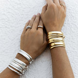 A close-up of arms wearing stacked Rendezvous Cuffs, Plus One Cuffs and All In Cuffs, one arm in silver and the other in gold, an Etched Paris Ring, a Tres Pearl Ring and a C'est Moi Link Ring against a textured white background.