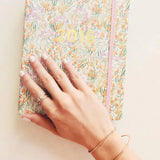 A hand wearing a Simple Bangle and a Diamond Cut Bracelet in rose gold and two rings, placed on top of a floral 2016 notebook.