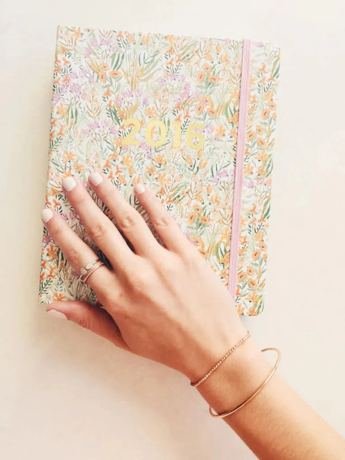 A hand wearing a Simple Bangle and a Diamond Cut Bracelet in rose gold and two rings, placed on top of a floral 2016 notebook.