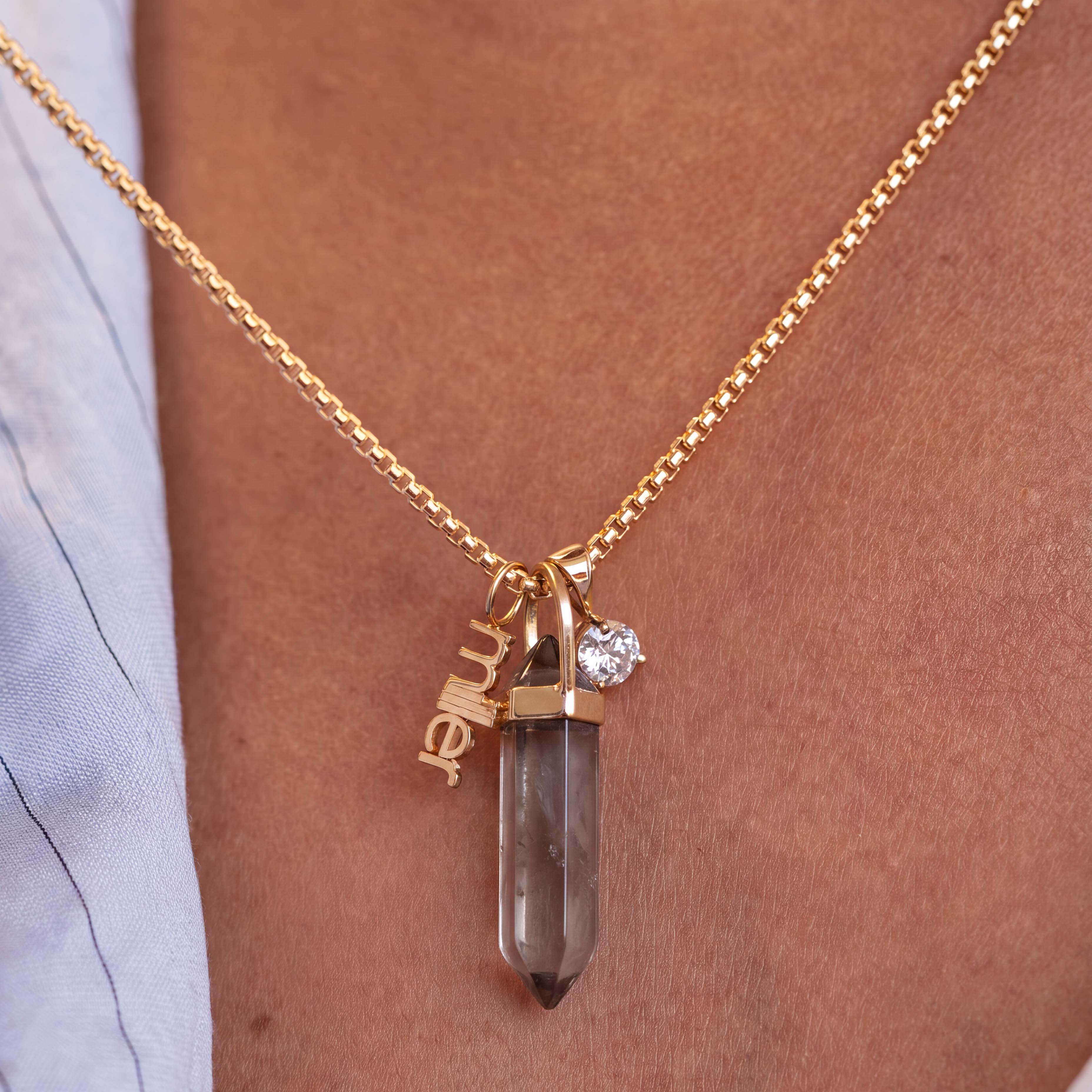 A gold chain necklace with a smoky quartz crystal pendant, featuring a small clear gemstone and a gold charm spelling 'miller', worn on a person's chest.