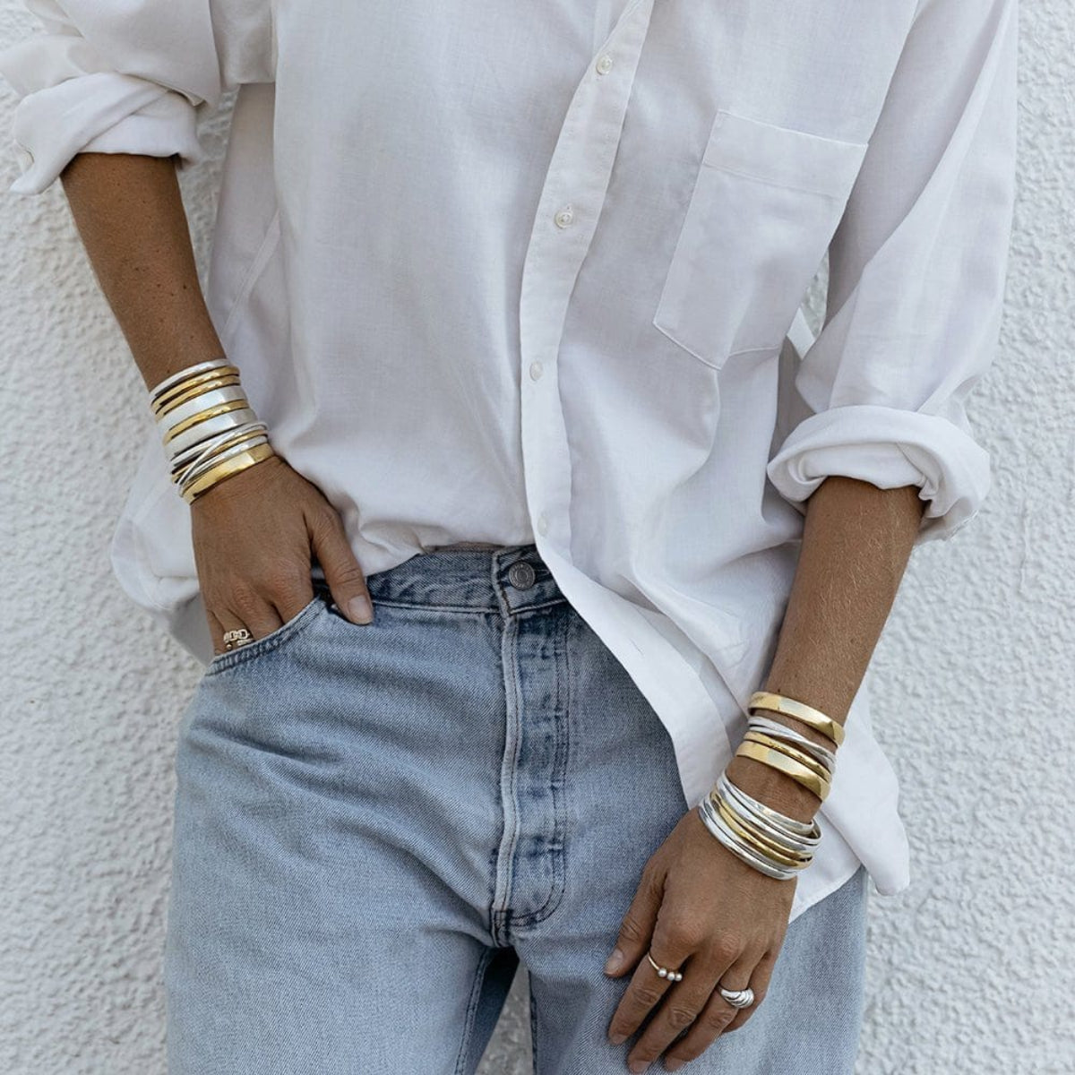 A person wearing silver and gold stacked Rendezvous Cuffs, Plus One Cuffs, All In Cuffs and Flirt Bangles on both arms, an Etched Paris Ring, a Tres Pearl Ring, a C’est Moi Link Ring and an Open Diamond Ring, styled in a white shirt and light blue jeans.