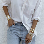 A person wearing silver and gold stacked Rendezvous Cuffs, Plus One Cuffs, All In Cuffs and Flirt Bangles on both arms, an Etched Paris Ring, a Tres Pearl Ring, a C’est Moi Link Ring and an Open Diamond Ring, styled in a white shirt and light blue jeans.