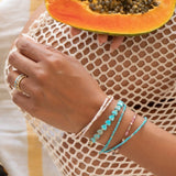 A hand wearing several Endless Summer Bracelets (Pink Sands, Turqs + Caicos, Coral Reef and Sea + Sky) and Bouquet Rings, holding a papaya in the other hand, wearing cream mesh clothing as the background.