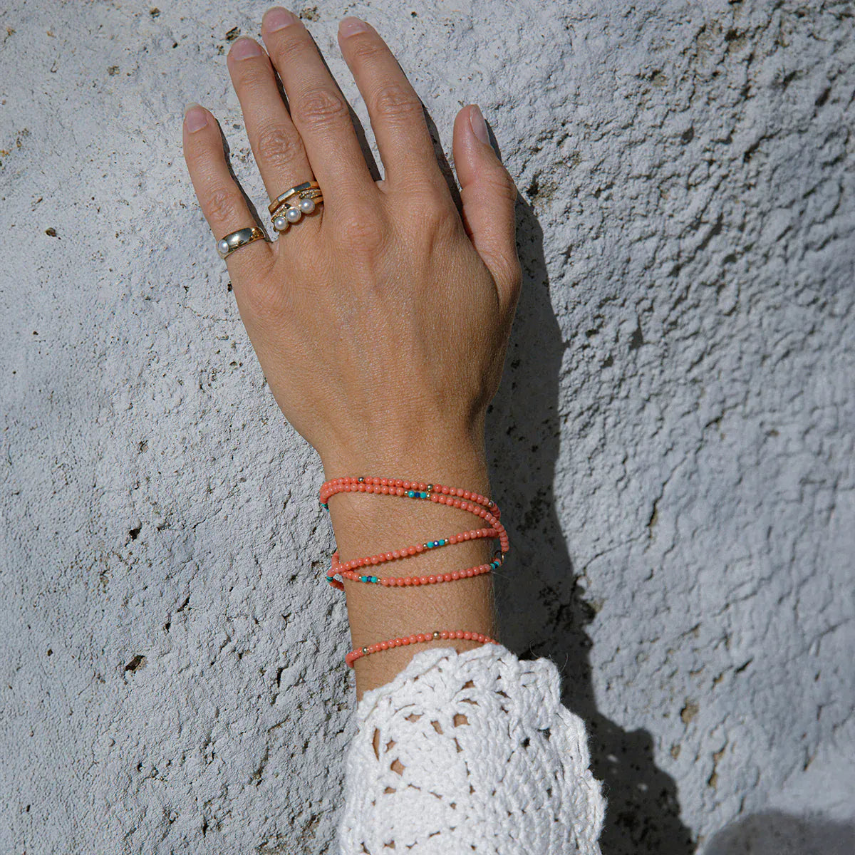 A hand wearing a Perla Pinky Ring, Tres Pearl Ring, an Open Diamond Ring, a Tenfold Ring and an Endless Summer Necklace - Calypso and an Endless Summer Bracelet - Mykonos with a light stone background. 